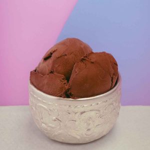 Small silver cup with a few scoops of rich brown chocolate ice cream. The cup sits on a texture white surface, against a split colour background of pink and purple