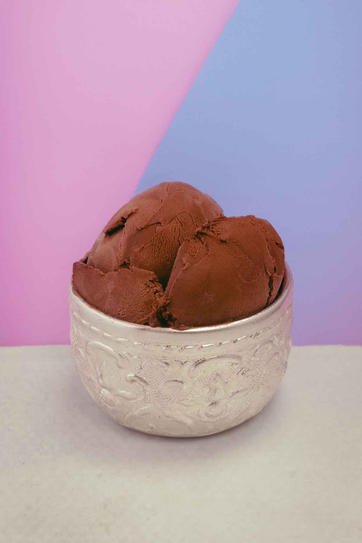 Small silver cup with a few scoops of rich brown chocolate ice cream. The cup sits on a texture white surface, against a split colour background of pink and purple