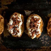 top down view of four twice baked potatoes on a well used oven tray. Two of the potatoes are topping with a fluffy white vegan cheese mixture and flecks of plant based bacon
