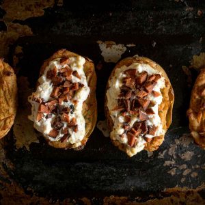 top down view of four twice baked potatoes on a well used oven tray. Two of the potatoes are topping with a fluffy white vegan cheese mixture and flecks of plant based bacon