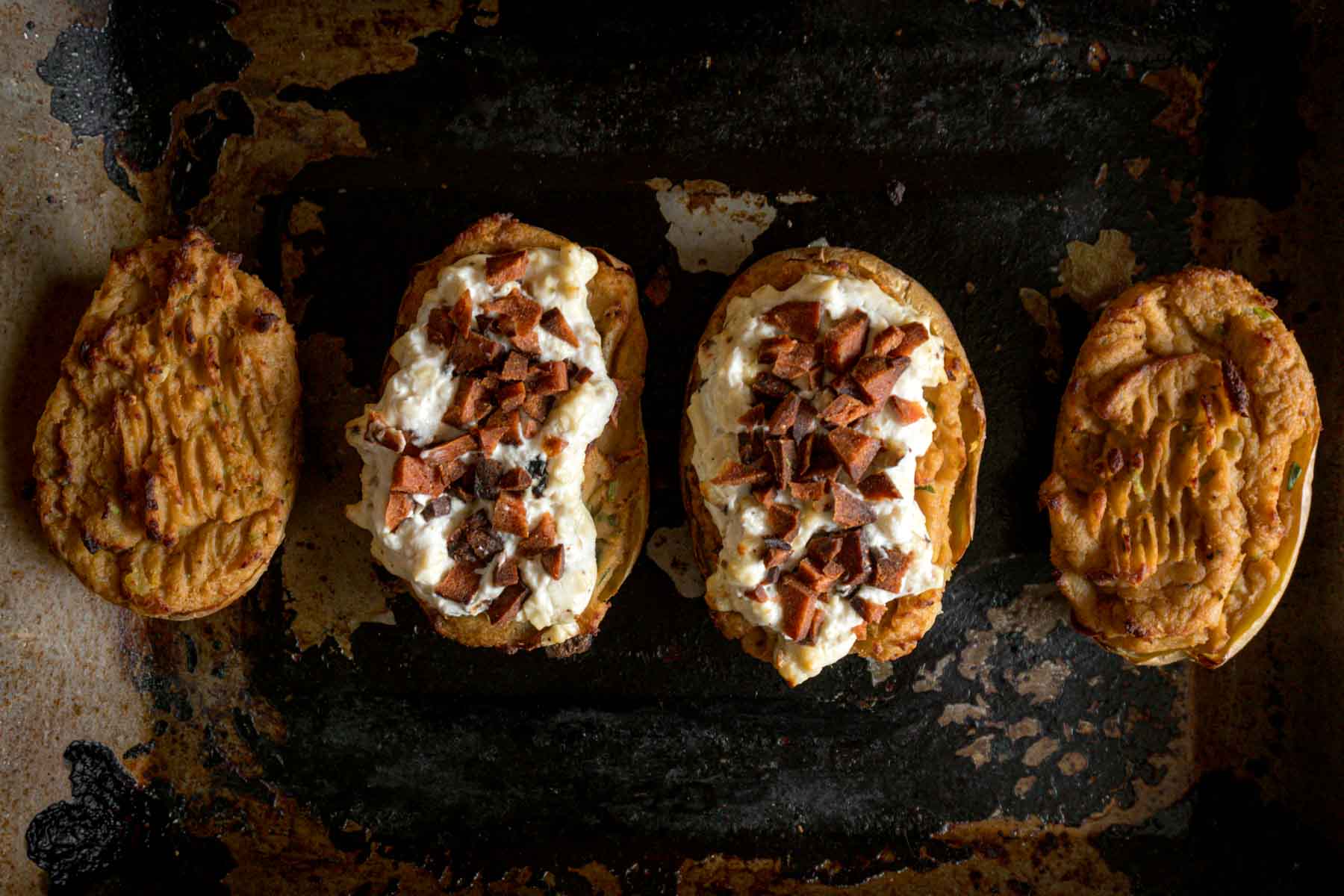 Vegan Twice Baked Potatoes top down view of four twice baked potatoes on a well used oven tray. Two of the potatoes are topping with a fluffy white vegan cheese mixture and flecks of plant based bacon