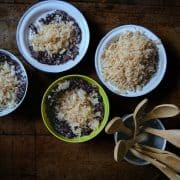 Top down view of four bowls, three with purple-black grains of rice with a caramelised coconut topping, and one with only coconut topping. A third metal container holds small wooden spoons