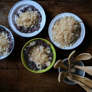Top down view of four bowls, three with purple-black grains of rice with a caramelised coconut topping, and one with only coconut topping. A third metal container holds small wooden spoons