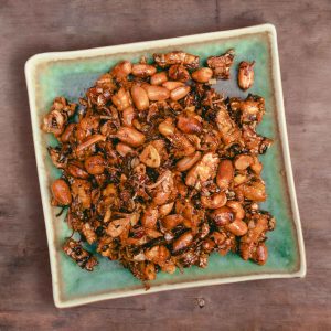 top down view of kering tempe. A blueish green square plate piled with shiny deep fried tempeh and peanuts