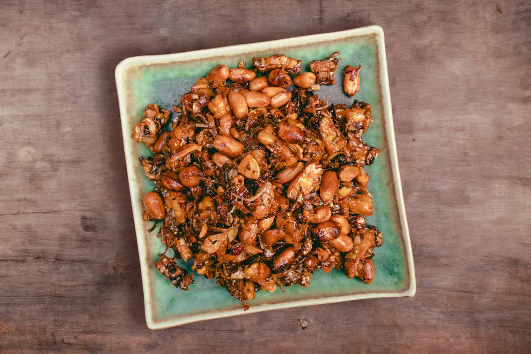 top down view of kering tempe. A blueish green square plate piled with shiny deep fried tempeh and peanuts