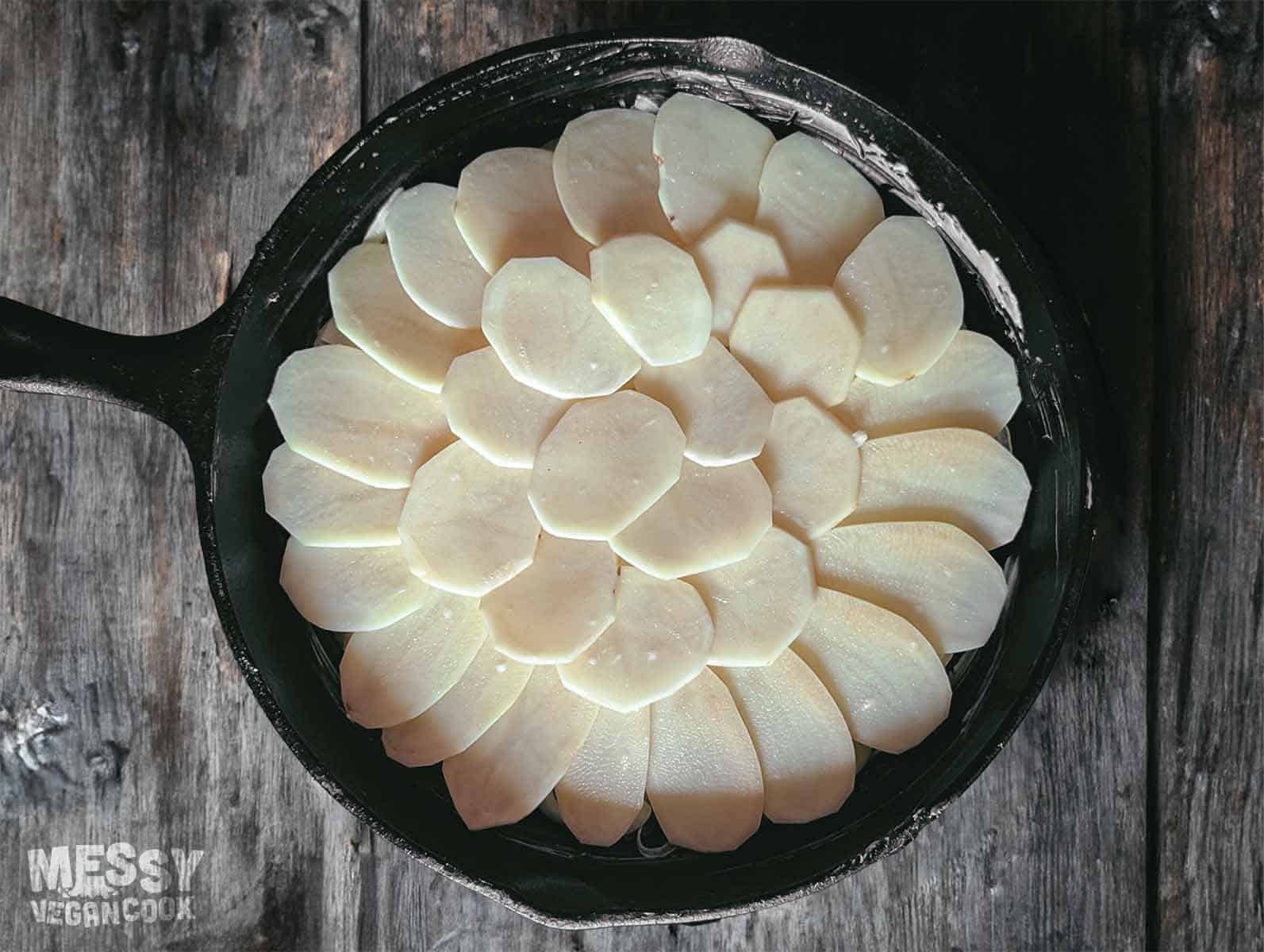 top down view of a cast iron skillet layered with thin slices of potato (uncooked)