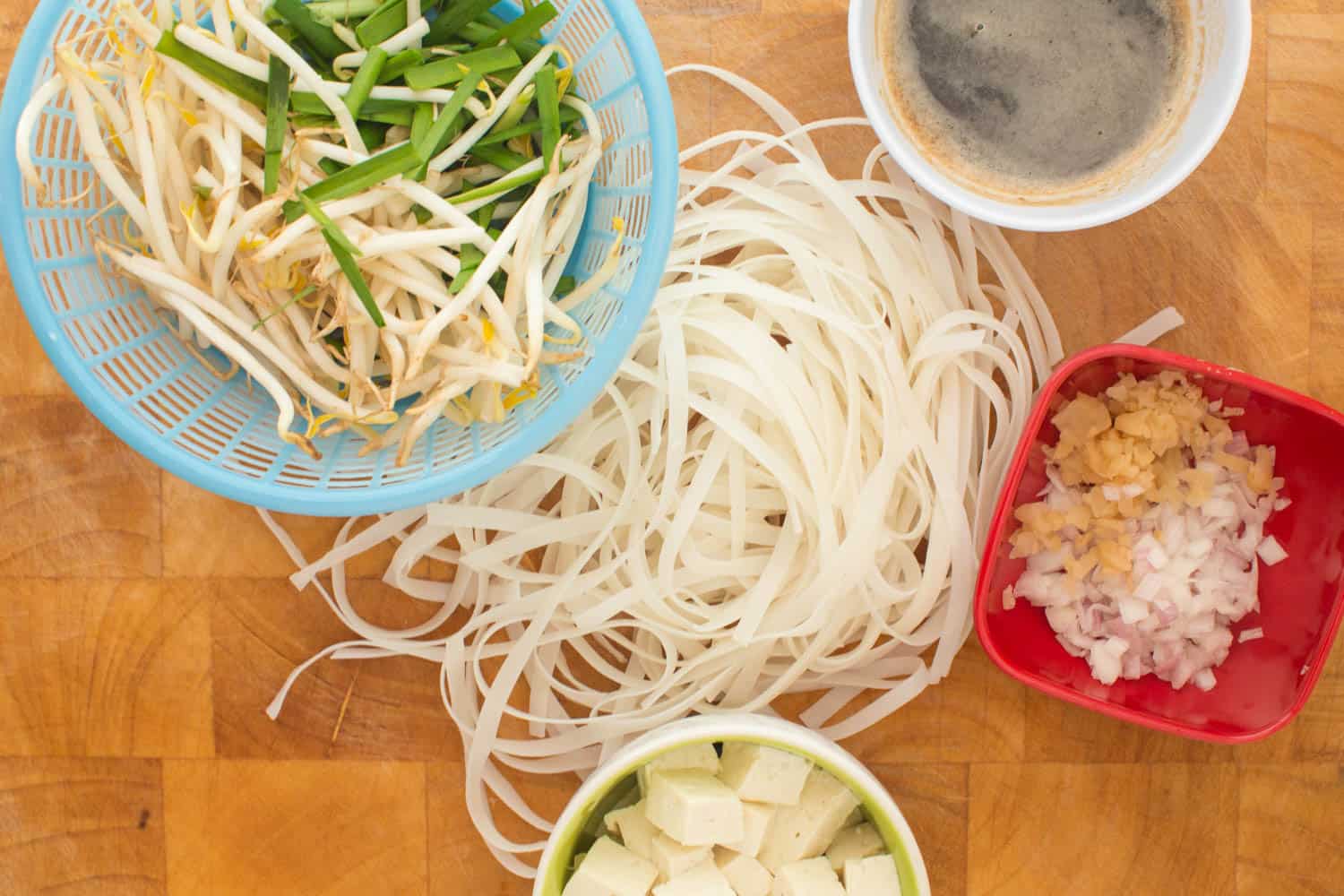 Pat thai ingredients laid out on a cutting board: Chinese chives, bean sprouts, tamarind sauce, preserved radish, shallots, tofu, rice noodles