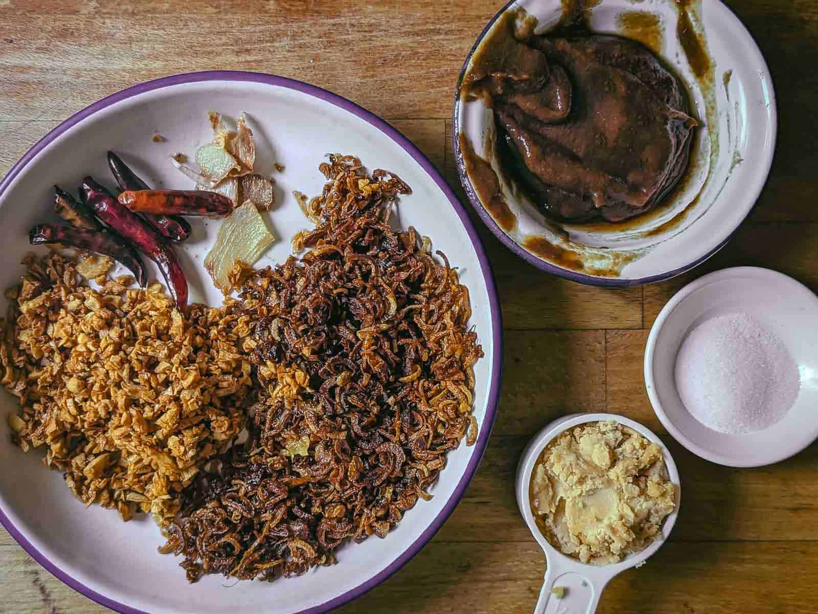 Top down image of all of the ingredients for making nam prik pao: a plate with fried shallots, fried garlic, fried galangal, and fried chillies; a measuring cup with palm sugar; a small bowl messily filled with dark brown tamarind paste; a tiny ramekin of salt