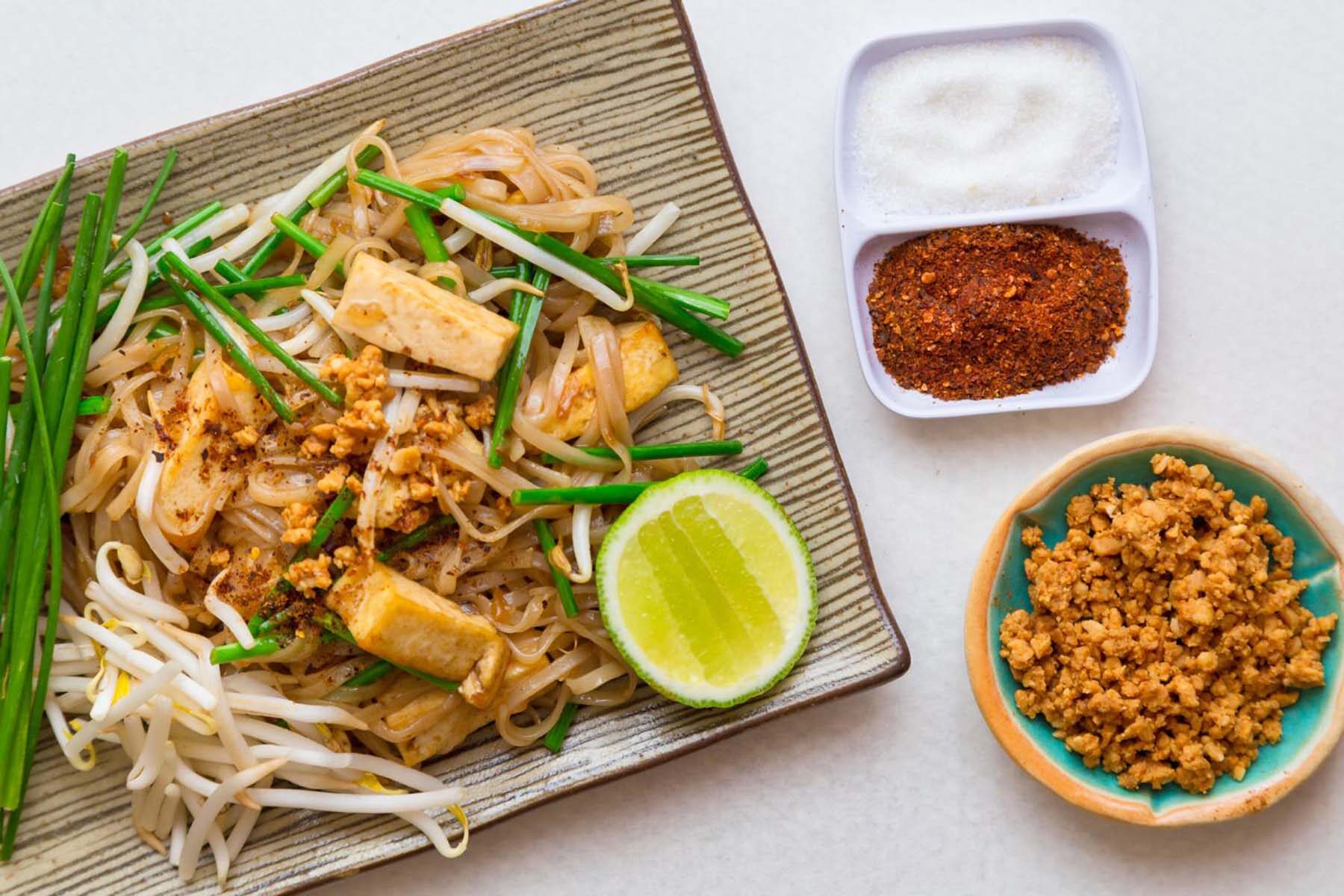 Top down view of vegan pad thai. Rectangular dish with a striped pattern, with a pile of noodles and green strips of garlic chives.