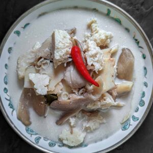 Black background with a bowl of tom kha soup on top. The bowl is shallow and features a silver rim and some blue-green pattern deeper in the bowl. The bowl contains white coconut based soup, with vegan chicken and oyster mushrooms piled up in the middle. There is a single small red chilli on top.