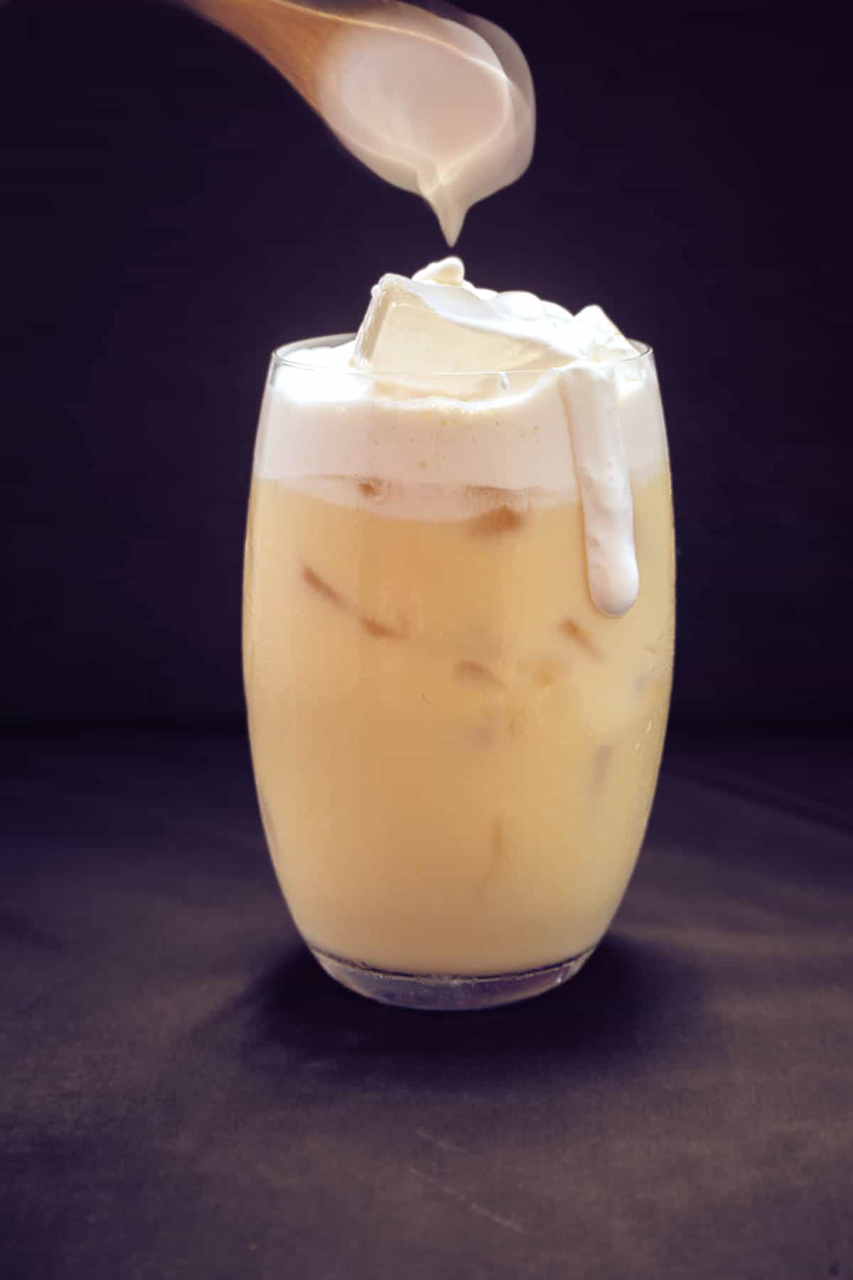 dark background with slightly curved glass filled ⅚ of the way with beige-yellow liquid (corn milk), topped with a creamy cheese foam (white). The foam is messily dripping down the side of the glass.