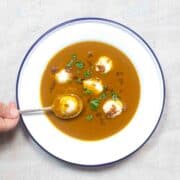 view from overhead of an enamelwear (white with blue rim) soup dish. Inside is orange soup with white blobs of peanut dumplings. A hand holds a spoon containing one dumpling.