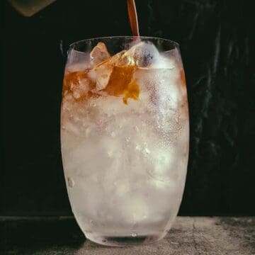 iced coconut water americano. Large glass filled with coconut water. Top left is a hand pouring espresso over the ice.