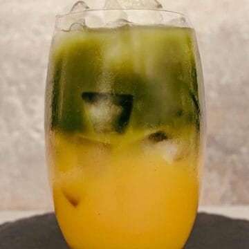 large glass with two layers of liquid. On the top is a deep green layer of matcha tea, and on the bottom a layer of orange juice.