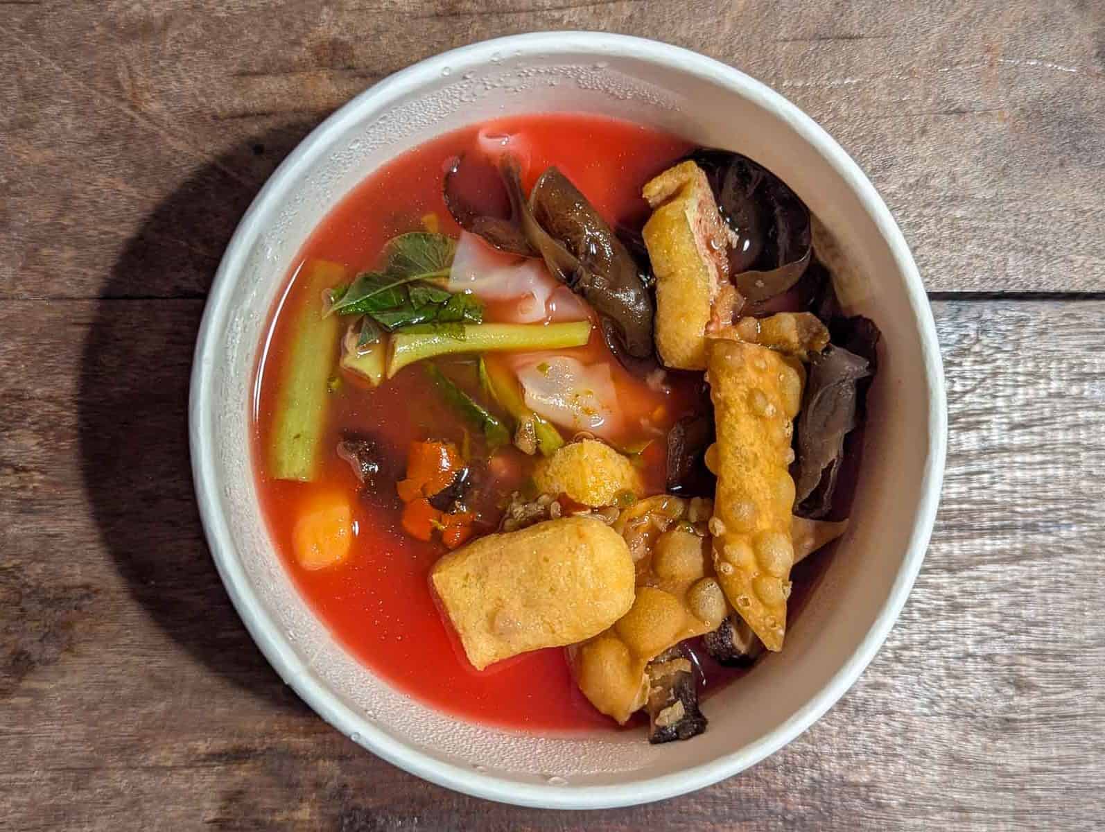 bright red thin soup in a cardboard bowl, including green vegetables, black mushrooms, and beige fried tofu bits