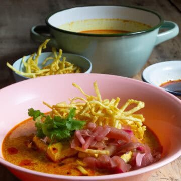 Pink plastic bowl of finished curry noodles with purple pickles, green herbs, and crispy yellow noodles.