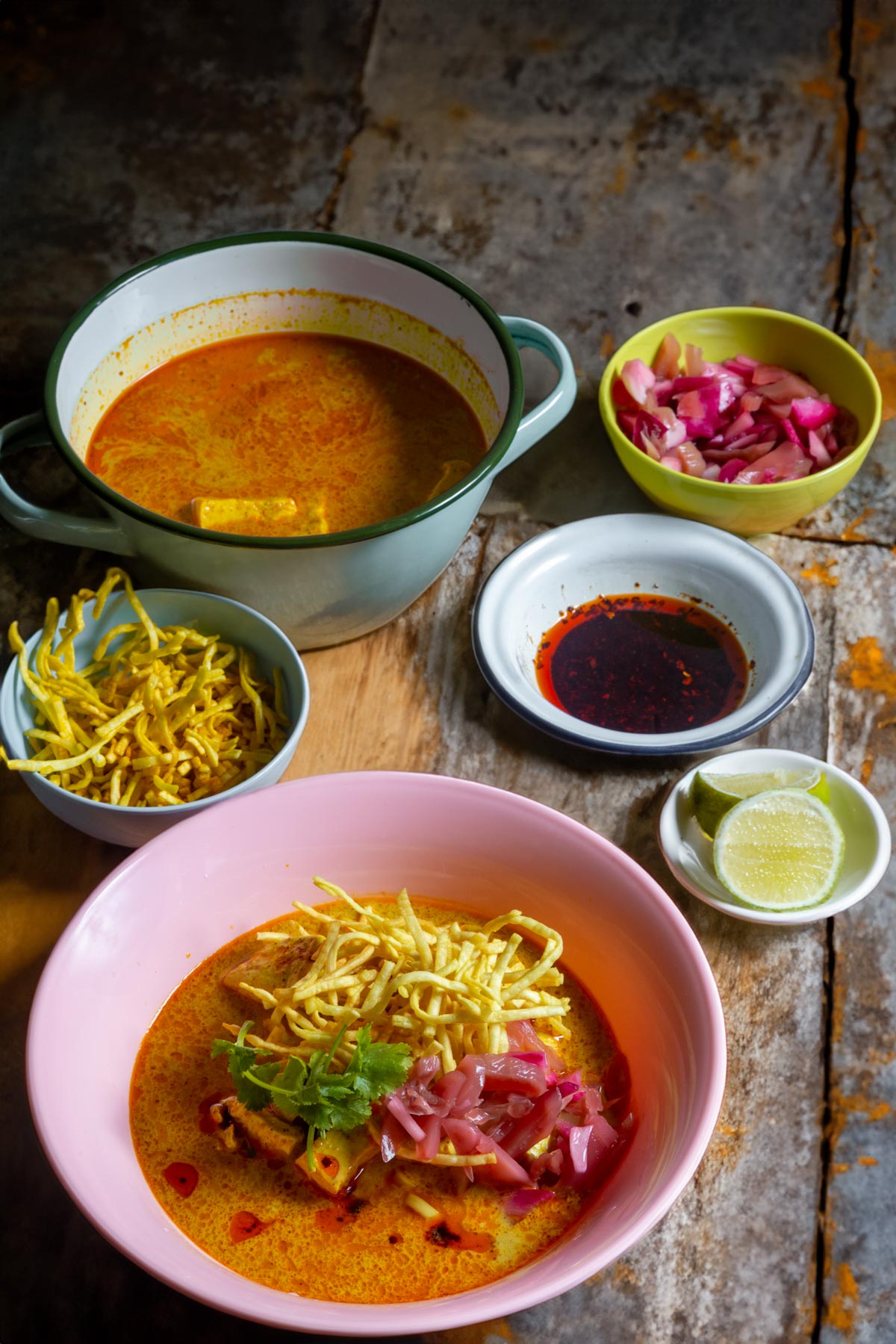 Image showcasing a bowl of prepared khao soi in a pink melamine bowl. Behind are a series of small dishes containing crispy fried noodles, lime wedges,, chilli oil, and pickles. At the back is a larger pot of khao soi curry.