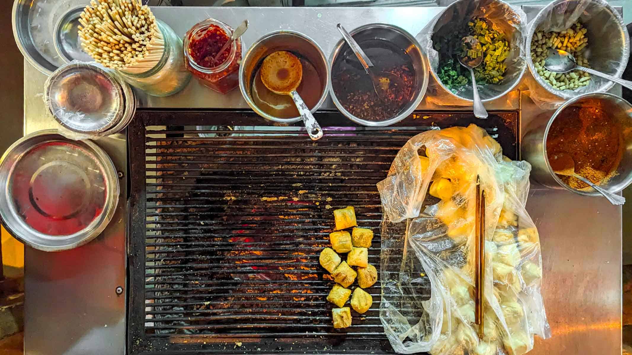 View from above of a rustic grill place with yellow cubes of tofu. The grill is surrounded by various mismatched jars and pots containing seasoning ingredients and chopsticks