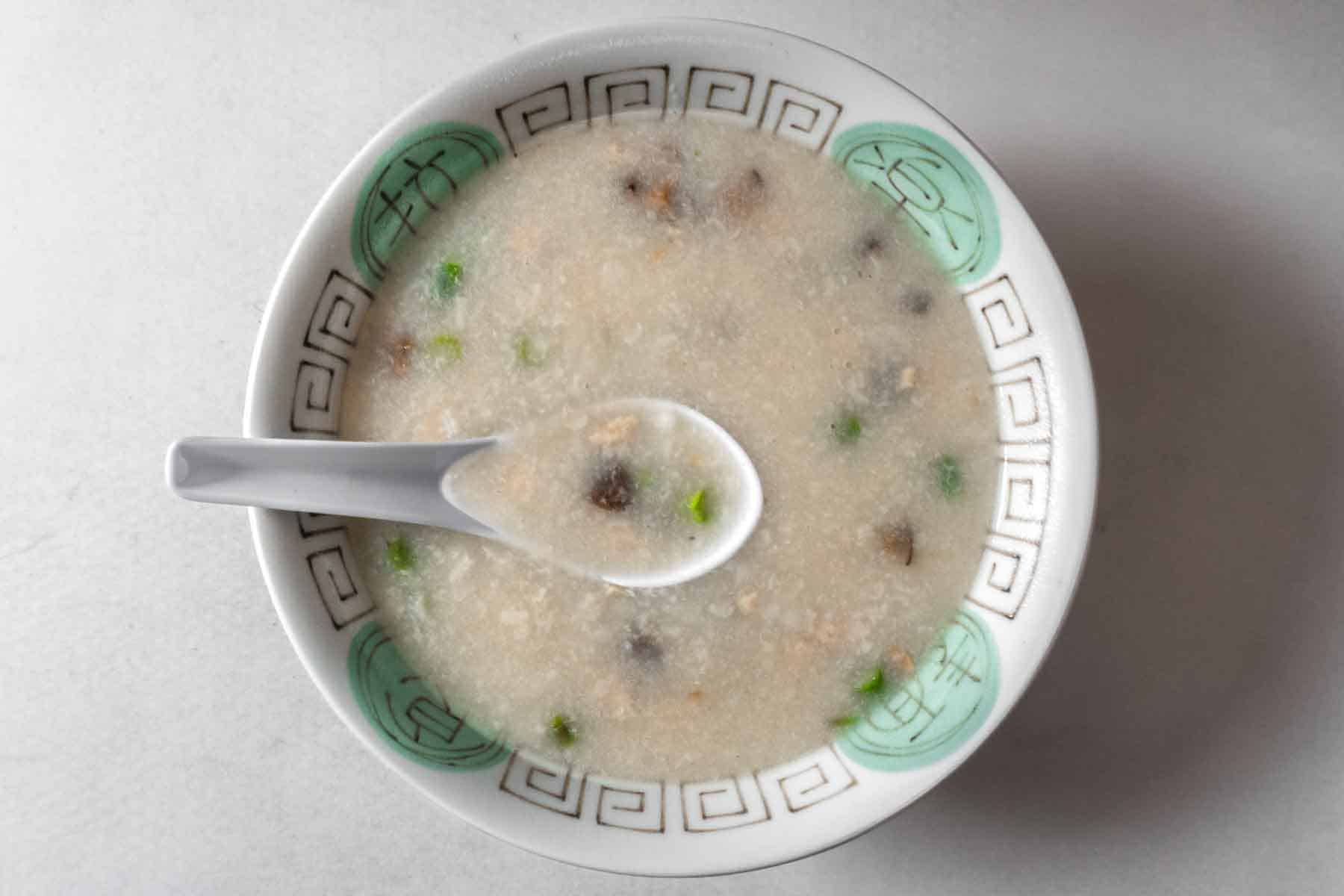 Top down view of vegan chicken congee in a patterned bowl with a Chinese soup spoon with congee in it