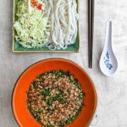 top down view of an orange bowl half filled with a soup topped with heaps of green herbs and chopped peanuts. Next to this is a plate with shredded white cabbage and rice noodles.
