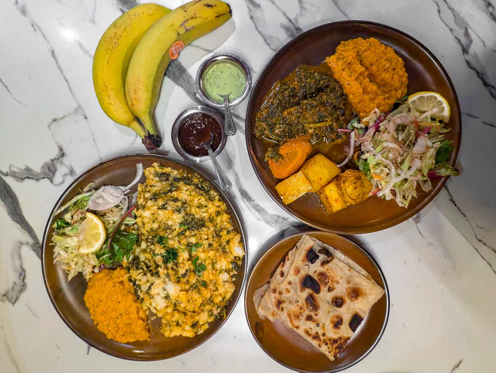 2 large brown plates containing a selection of stews and salads; one small plate with squares of flatbread, 2 ramekins with chutney, and two unpeeled bananas on a white marble surface