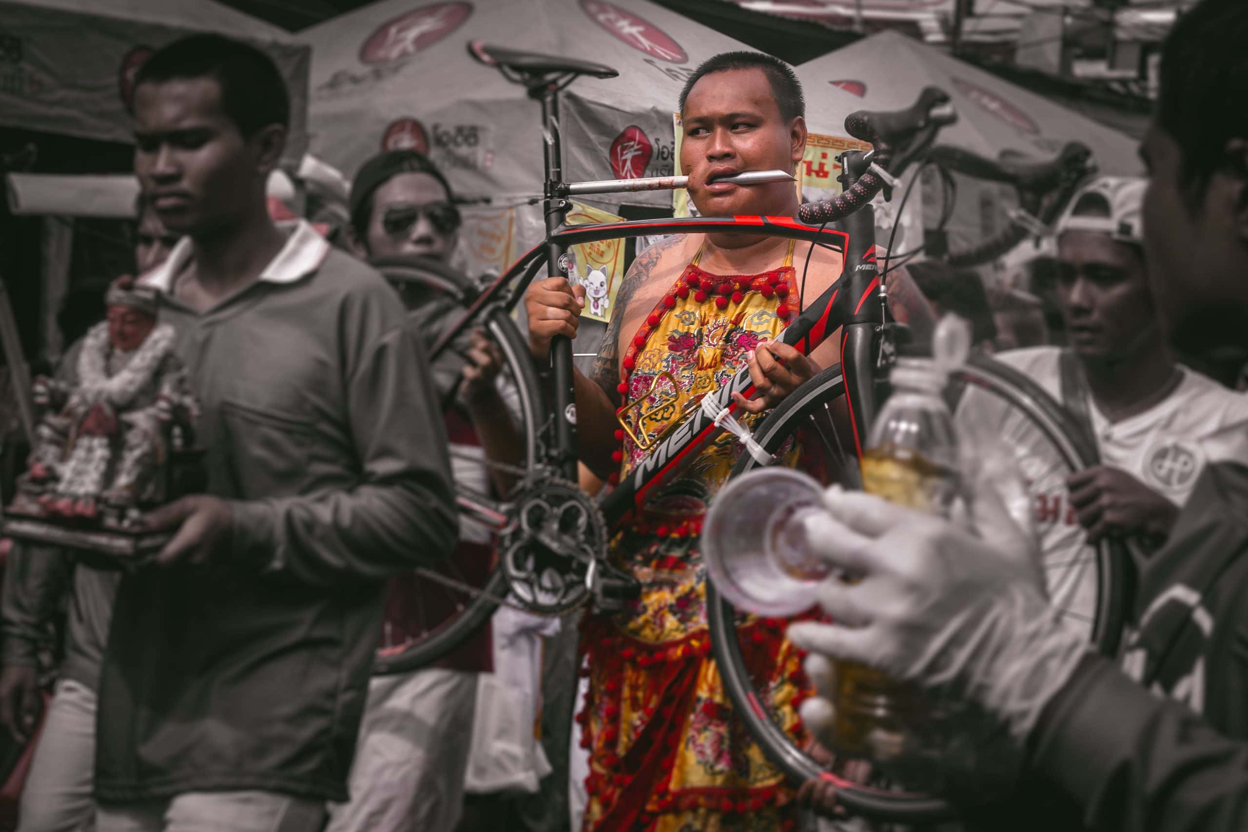 Spirit medium with a red apron. His cheek is pierced with the central bar of a bicycle, while he carries the bike with both hands
