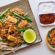 Top down view of vegan pad thai. Rectangular dish with a striped pattern, with a pile of noodles and green strips of garlic chives.