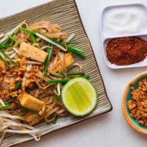 Top down view of vegan pad thai. Rectangular dish with a striped pattern, with a pile of noodles and green strips of garlic chives.