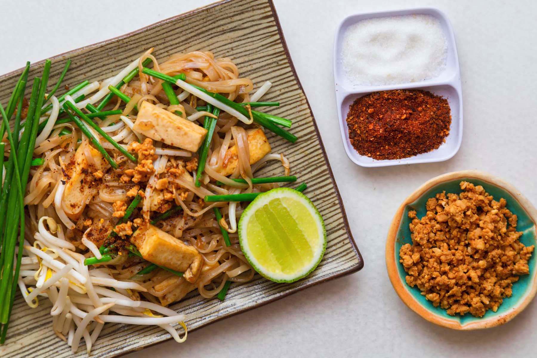 Top down view of vegan pad thai. Rectangular dish with a striped pattern, with a pile of noodles and green strips of garlic chives.