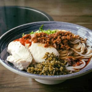Blue and white ceramic bowl filled with chunky round noodles in a small amount of red broth. Finely chopped green-yellow pickled mustard greens, large spoonfuls of tofu, bright red chilli sauce, and a vegan meat mixture top the noodles.