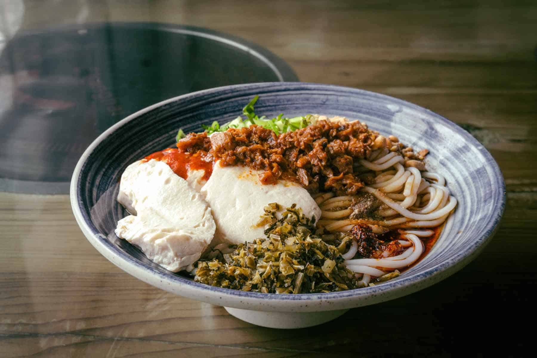 Blue and white ceramic bowl filled with chunky round noodles in a small amount of red broth. Finely chopped green-yellow pickled mustard greens, large spoonfuls of tofu, bright red chilli sauce, and a vegan meat mixture top the noodles.